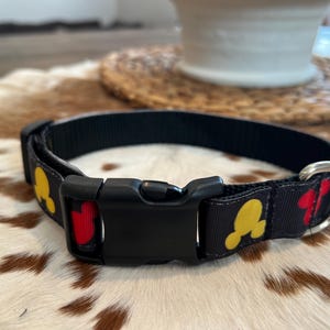 Könnte beinhalten: Schwarzes Hundehalsband mit einem sich wiederholenden Muster aus gelben und roten Zeichentrickfigurenköpfen. Das Halsband hat eine schwarze Kunststoffschnalle und einen silbernen Metall-D-Ring. Das Halsband wird auf einer braun-weiß gefleckten Oberfläche präsentiert.