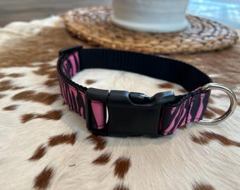 Könnte beinhalten: Halsband für Hunde mit rosa und schwarzem Zebra-Print. Es hat eine schwarze Kunststoffschnalle und einen silbernen Metall-D-Ring. Das Halsband ist aus Nylon gefertigt. Ein modisches Accessoire für Haustiere.