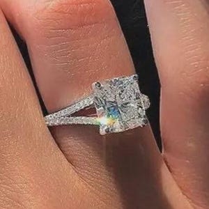 May include: A silver-toned ring featuring a large, rectangular radiant-cut center stone with a faceted surface. The band is split into two thin, pavé-set arms that meet at the base, creating a delicate, multi-faceted appearance on the finger.