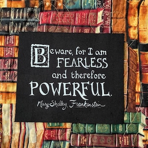 May include: A black square with white text quoting Mary Shelley's Frankenstein, set against a backdrop of fabric printed with images of old books. The quote reads: "Beware, for I am FEARLESS and therefore POWERFUL."