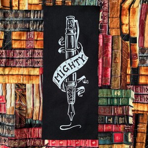 May include: A black rectangular fabric panel featuring a white illustration of a fountain pen with the word "MIGHTY" on a banner. The background is a patchwork of various antique books in shades of brown, red, and green.