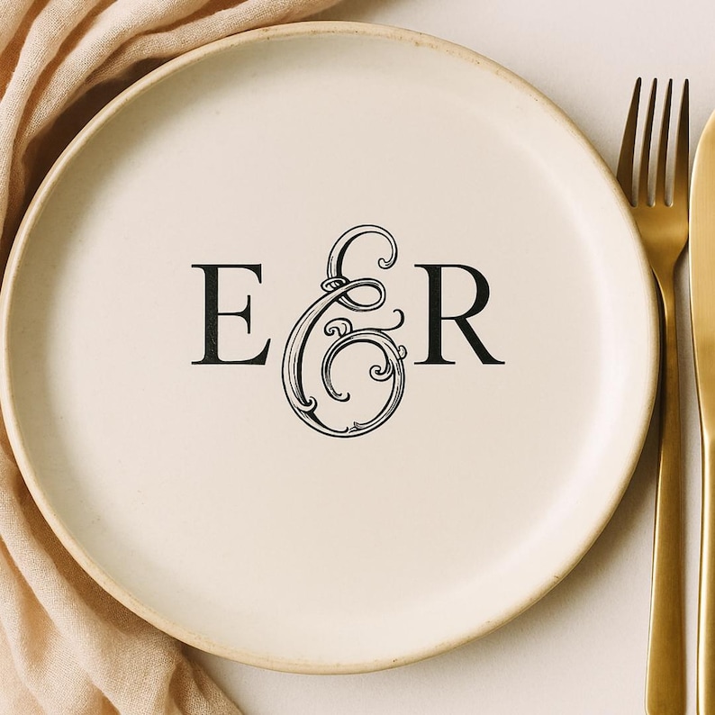 May include: A cream-colored ceramic plate with the black letters "E & R" in a decorative font. A gold fork and knife are to the right of the plate. A beige cloth is draped around the plate.