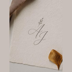 May include: A close-up of a textured, off-white paper with the intertwined letters "A" and "J" in a delicate, cursive script. A small sprig of leaves sits above the letters. A dried, brown leaf rests on the paper, and a brown fabric is visible in the upper left corner.