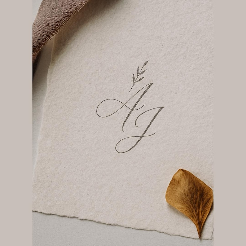 May include: A close-up of a textured, off-white paper with the intertwined letters "A" and "J" in a delicate, cursive script. A small sprig of leaves sits above the letters. A dried, brown leaf rests on the paper, and a brown fabric is visible in the upper left corner.