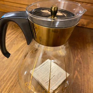 May include: Vintage Pyrex coffee pot with a clear glass body, black handle, and gold-colored accents. The lid is black with a gold knob. The glass carafe has gold decorative lines and a paper insert. A classic kitchenware item.