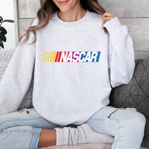 May include: A light gray sweatshirt featuring the word "NASCAR" in a colorful, horizontal design across the chest. The word is in a bold, sans-serif font with a gradient of yellow, red, pink, and blue.