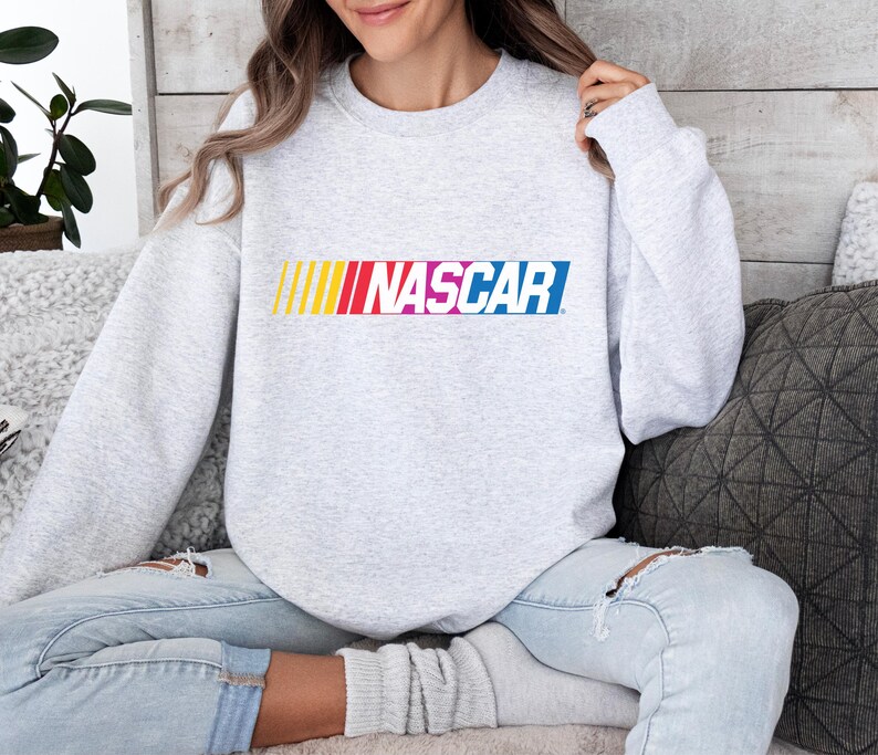 May include: A light gray sweatshirt featuring the word "NASCAR" in a colorful, horizontal design across the chest. The word is in a bold, sans-serif font with a gradient of yellow, red, pink, and blue.