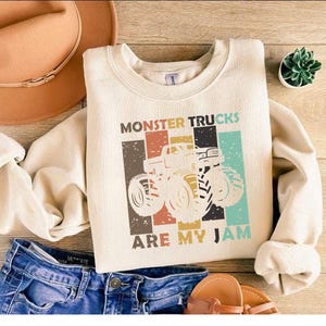 May include: Cream-colored sweatshirt with the text "MONSTER TRUCKS ARE MY JAM" and a graphic of a monster truck. The sweatshirt is paired with blue denim shorts, a brown hat, and sandals. A small succulent plant is also visible.