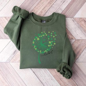 May include: A green sweatshirt featuring a dandelion design with shamrock accents. The word "lucky" is written in script below the design. The sweatshirt is made of a soft material and has ribbed cuffs and a crew neck.