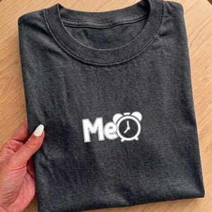 May include: Dark gray t-shirt with a white graphic that says "Me" with a clock icon. The shirt is folded neatly on a wooden surface.