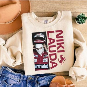 May include: Cream-colored sweatshirt featuring a graphic of Niki Lauda in a racing suit and helmet. The text "NIKI LAUDA" is printed in red, along with a Ferrari logo. The sweatshirt is paired with blue jeans, a brown hat, and sandals.
