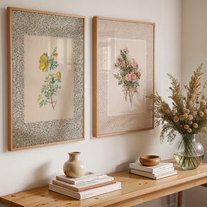 May include: Two framed botanical prints with floral illustrations. One shows yellow flowers, the other pink. The frames are light wood with patterned borders. A wooden table displays books, a vase, and a bowl.