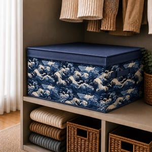 May include: A rectangular storage box with a solid dark blue lid and a body featuring a repeating pattern of white stylised ocean waves on a dark blue background. The box includes integrated side handles and sits on a shelf in a closet space.