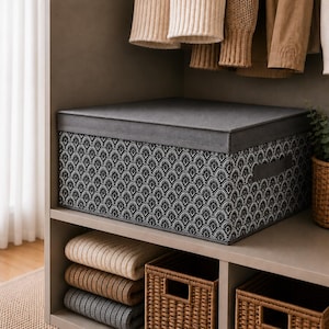 May include: A grey storage box with a patterned design, on a shelf. The box has a grey lid and handles. Below the box are folded jumpers and woven baskets. The background includes hanging clothes and a potted plant.