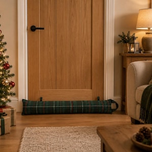 May include: A long, cylindrical door draught excluder featuring a dark green, blue, and red plaid tartan pattern. The fabric accessory includes two small fabric loops at the top and a handle on the right side, positioned horizontally at the base of a door.
