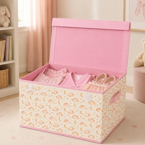 May include: A pink storage box with a lid, containing pink clothes and shoes. The box features a cream background with a pattern of clouds and moons. The interior and trim are pink.