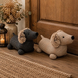May include: Two dog-shaped doorstops made of fabric with a plaid pattern. One doorstop is dark grey and black, while the other is light beige and brown. Both feature floppy ears with a fringed texture, a small tail, and a black nose.