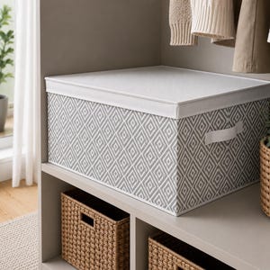 May include: A grey and white patterned storage box with a white lid and handle. The rectangular box sits on a shelf above two woven baskets. Ideal for storing items, the box features a geometric design.