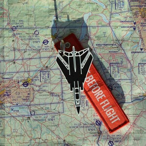 Peut inclure: Un porte-clés noir en forme de jet et une étiquette rouge avec le texte "BEFORE FLIGHT" sont posés sur une carte aéronautique détaillée. La carte présente des informations géographiques et de navigation.