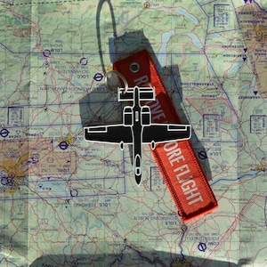 May include: A black and white airplane keychain with a red "REMOVE BEFORE FLIGHT" tag, resting on a detailed map. The keychain is a silhouette of a military aircraft.