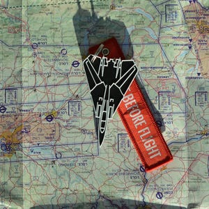 May include: A black and white fighter jet keychain and a red "BEFORE FLIGHT" tag rest on a detailed map. The keychain features a stylized jet design. The tag is rectangular.