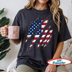 Op de afbeelding: Een antracietgrijs T-shirt met korte mouwen en een afbeelding van de Amerikaanse vlag die door de stof lijkt te scheuren. Het T-shirt wordt gecombineerd met een lichtroze mok versierd met witte stippen en gouden cursieve tekst.
