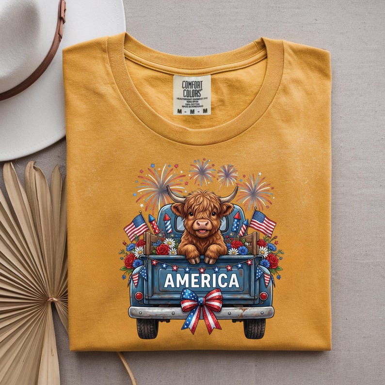 May include: Mustard yellow t-shirt featuring a cartoon Highland cow in a blue pickup truck with the word "AMERICA". The truck is decorated with American flags, fireworks, and a red, white, and blue bow.