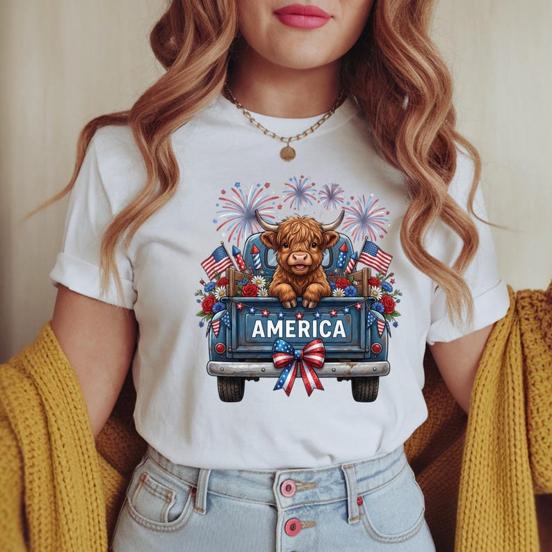 May include: White t-shirt featuring a graphic of a Highland cow in a blue pickup truck bed decorated with American flags, flowers, and fireworks. The truck bed has the word "AMERICA" on it. A red, white, and blue bow is at the bottom.