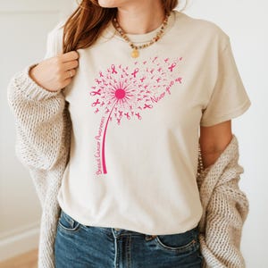 Op de afbeelding: Een beige t-shirt met korte mouwen en een roze afbeelding van een paardenbloem waarvan de zaden kleine lintjes voor borstkankerbewustzijn zijn. De tekst "Breast Cancer Awareness" staat langs de steel en "Never give up" staat rechts.