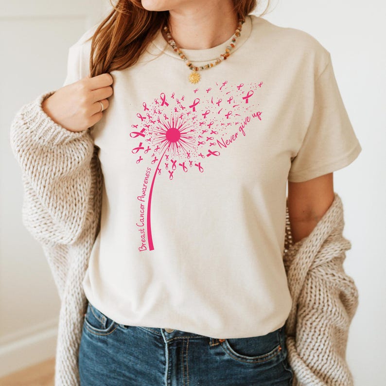 Op de afbeelding: Een beige t-shirt met korte mouwen en een roze afbeelding van een paardenbloem waarvan de zaden kleine lintjes voor borstkankerbewustzijn zijn. De tekst "Breast Cancer Awareness" staat langs de steel en "Never give up" staat rechts.