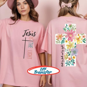 May include: Pink t-shirts with the text "Jesus is the Light" and a cross graphic. The back of the shirt features a floral cross design with the text "Walk by faith not by sight". The shirts are a light pink color.