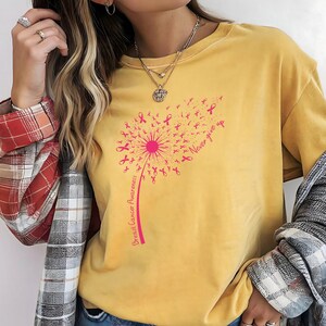 Op de afbeelding: Een mosterdgeel t-shirt met een roze afbeelding van een paardenbloem waarvan de zaden kleine lintjes voor borstkankerbewustzijn zijn. De tekst "Breast Cancer Awareness" staat langs de steel en "Never give up" staat rechtsboven gedrukt.
