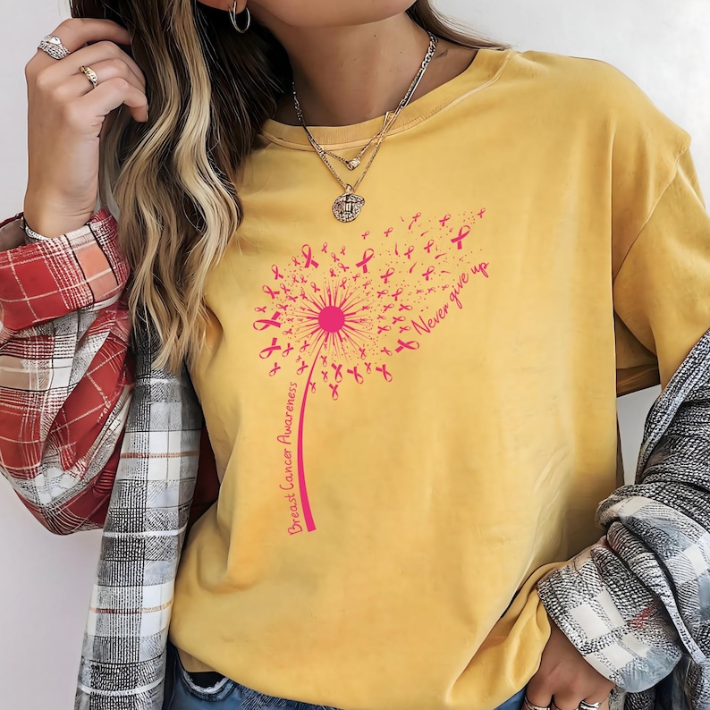 Op de afbeelding: Een mosterdgeel t-shirt met een roze afbeelding van een paardenbloem waarvan de zaden kleine lintjes voor borstkankerbewustzijn zijn. De tekst "Breast Cancer Awareness" staat langs de steel en "Never give up" staat rechtsboven gedrukt.