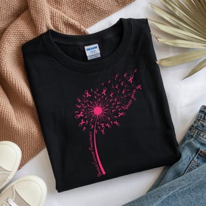Op de afbeelding: Een zwart T-shirt met ronde hals en een felroze afbeelding van een paardenbloem waarvan de zaden kleine lintjes voor borstkankerbewustzijn zijn. De tekst "Never give up" en "Breast Cancer Awareness" is in het roze gedrukt.