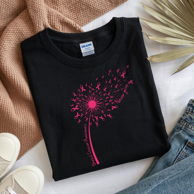 Op de afbeelding: Een zwart T-shirt met ronde hals en een felroze afbeelding van een paardenbloem waarvan de zaden kleine lintjes voor borstkankerbewustzijn zijn. De tekst "Never give up" en "Breast Cancer Awareness" is in het roze gedrukt.