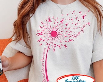 May include: A light heather gray t-shirt featuring a pink graphic of a dandelion where the seeds are small breast cancer awareness ribbons. The text "Breast Cancer Awareness" runs along the stem, and "Never give up" is printed to the right.