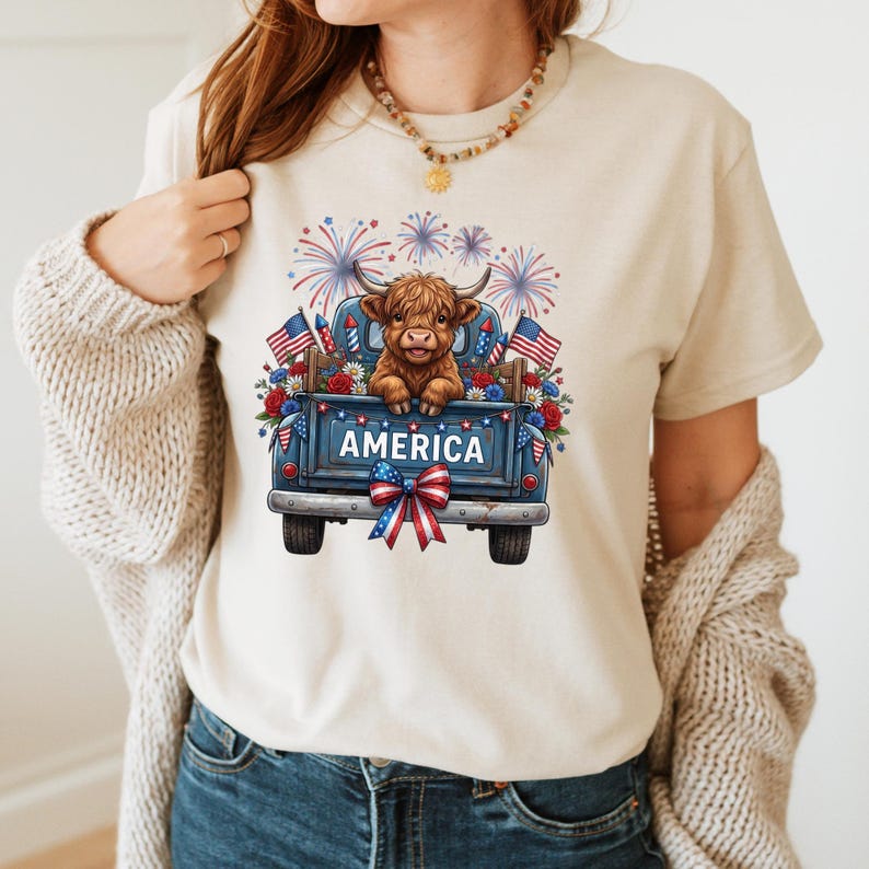 May include: Beige t-shirt featuring a blue truck bed graphic with the word "AMERICA". The truck bed is filled with flowers, flags, and a Highland cow. Fireworks are displayed in the background.