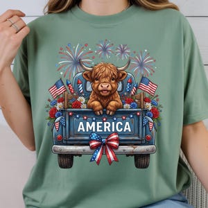 May include: A sage green t-shirt featuring a graphic of a Highland cow in a blue truck bed decorated with American flags, fireworks, and the word "AMERICA". The design has a patriotic theme.