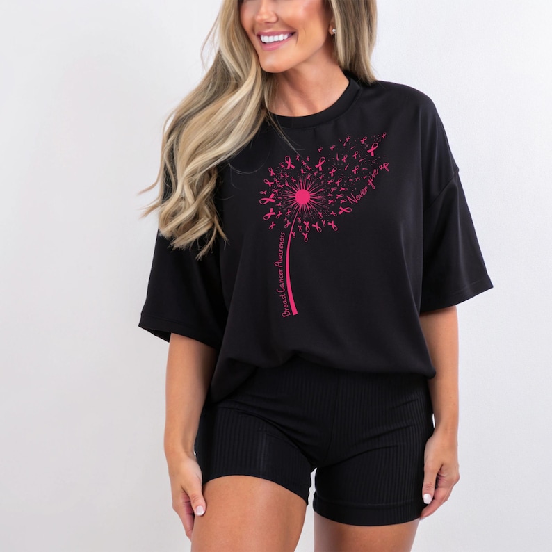 Op de afbeelding: Een zwart T-shirt met korte mouwen en een roze afbeelding van een paardenbloem gemaakt van lintjes voor borstkankerbewustzijn. De steel bevat de tekst Breast Cancer Awareness en de zin Never give up in roze letters.