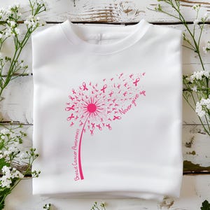 Op de afbeelding: Een wit t-shirt met een roze afbeelding van een paardenbloem waarvan de zaden kleine lintjes voor borstkankerbewustzijn zijn. De steel bevat de tekst Breast Cancer Awareness en de zin Never give up in een roze cursief lettertype.