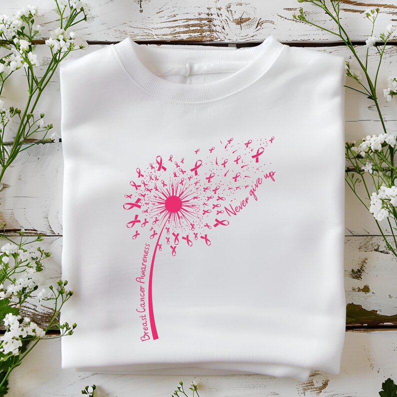 Op de afbeelding: Een wit t-shirt met een roze afbeelding van een paardenbloem waarvan de zaden kleine lintjes voor borstkankerbewustzijn zijn. De steel bevat de tekst Breast Cancer Awareness en de zin Never give up in een roze cursief lettertype.