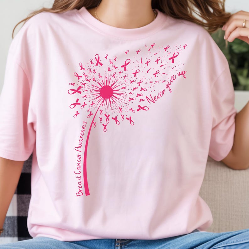 Op de afbeelding: Een lichtroze t-shirt met een donkerroze afbeelding van een paardenbloem waarvan de zaden kleine lintjes voor borstkankerbewustzijn zijn. De tekst "Breast Cancer Awareness" staat op de steel en "Never give up" is in cursief geschreven.