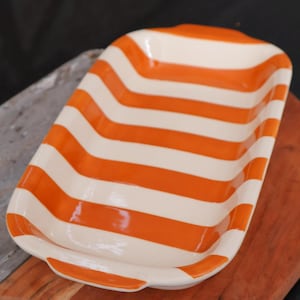 Könnte beinhalten: Rechteckige Servierschale aus Keramik mit horizontalen orangefarbenen und weißen Streifen. Die Schale hat eine leicht gebogene Form mit kleinen Griffen an jedem Ende. Sie liegt auf einem Holzschneidebrett.