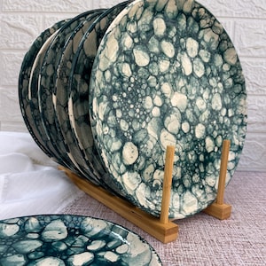 Könnte beinhalten: Ein Set aus sechs Keramikplatten mit einem einzigartigen Design. Die Teller weisen ein türkis-weißes Muster auf, das an organische Formen erinnert. Sie werden in einem Holzplattenständer präsentiert, mit einem Teller separat. Eine dekorative Ergänzung für jede Küche.