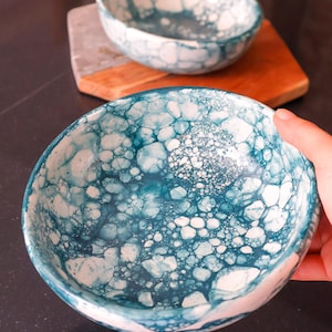 Op de afbeelding: Twee keramische kommen met een uniek blauw en wit bubbelpatroon. De kommen staan op een houten snijplank en een donker oppervlak. Ideaal om eten in te serveren of als decoratieve stukken.