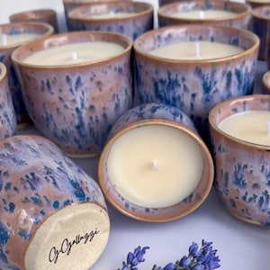 Könnte beinhalten: Handgefertigte Kerzen in Keramikbehältern mit einer gesprenkelten blauen und rosa Glasur. Jede Kerze hat eine weiße Wachsfüllung und einen einzelnen Docht. Ein Zweig Lavendel ist im Vordergrund. Signatur des Künstlers auf einem Topf.