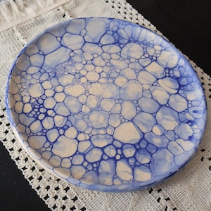 Op de afbeelding: Een ronde keramische plaat met een blauw en wit bellenpatroon. De plaat ligt op een wit kanten kleedje. Het ontwerp van de plaat toont een netwerk van onderling verbonden vormen, wat een unieke en artistieke uitstraling creëert.