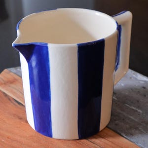 Può includere: Brocca in ceramica bianca con strisce blu verticali. La brocca ha un beccuccio e un manico. Il design è semplice e classico, adatto per servire bevande. La brocca è appoggiata su un tagliere di legno.