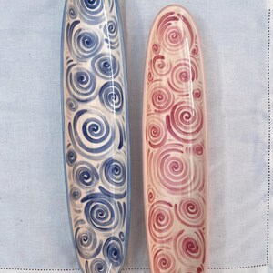 May include: Two ceramic serving platters. One is blue with a white interior and blue spiral design. The other is pink with a pink spiral design. Both are oval-shaped with a raised edge.