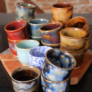 Könnte beinhalten: Eine Sammlung handgefertigter Keramikbecher in verschiedenen Farben, darunter Blau, Rot und Braun. Die Becher haben eine einzigartige, glasierte Oberfläche und sind auf einer Holzoberfläche gestapelt. Die Becher sind etwa 7.6 cm hoch.